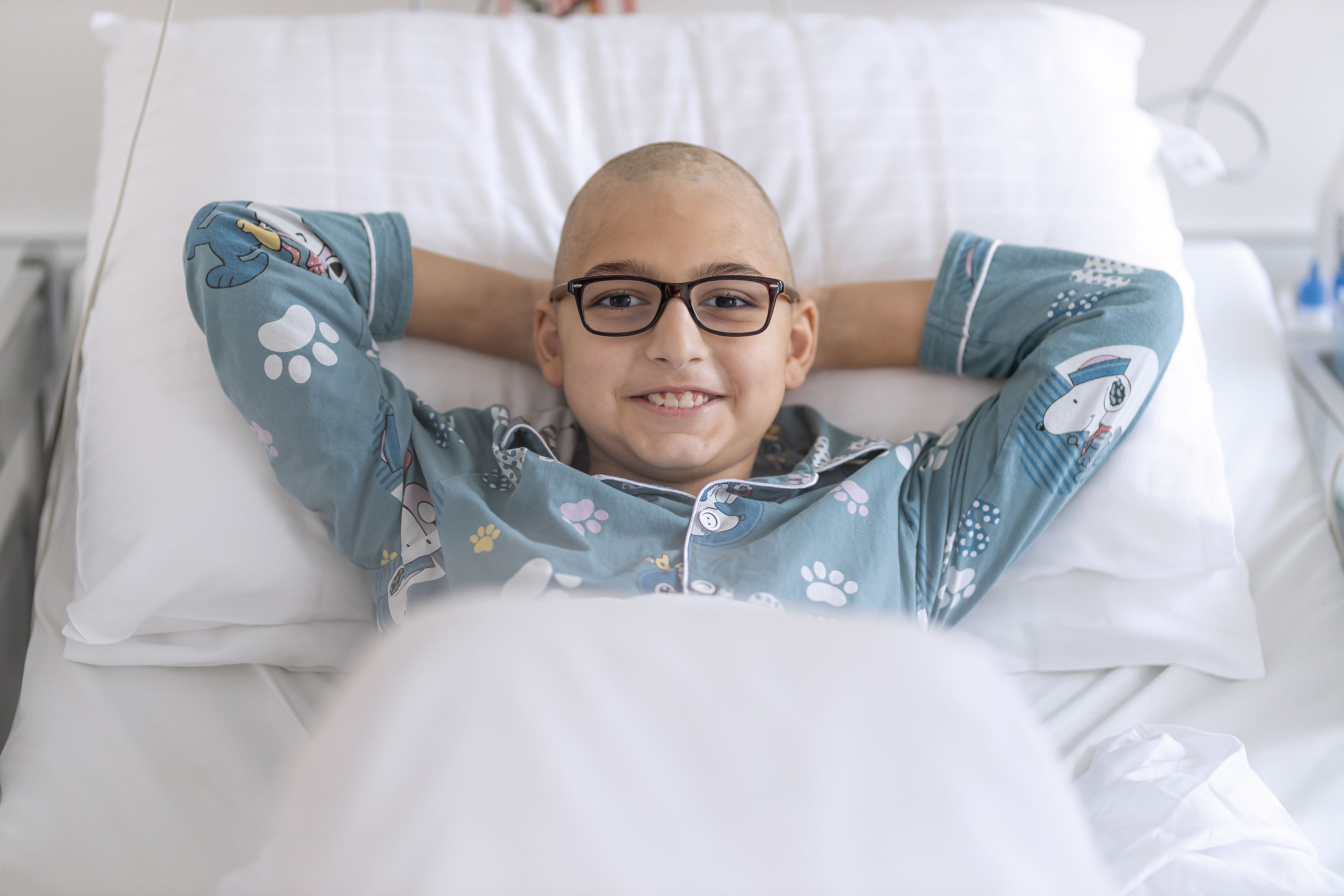 Ein lächelnder junger Mensch mit Brille und Schlafanzug liegt mit den Händen hinter dem Kopf in einem Krankenhausbett.