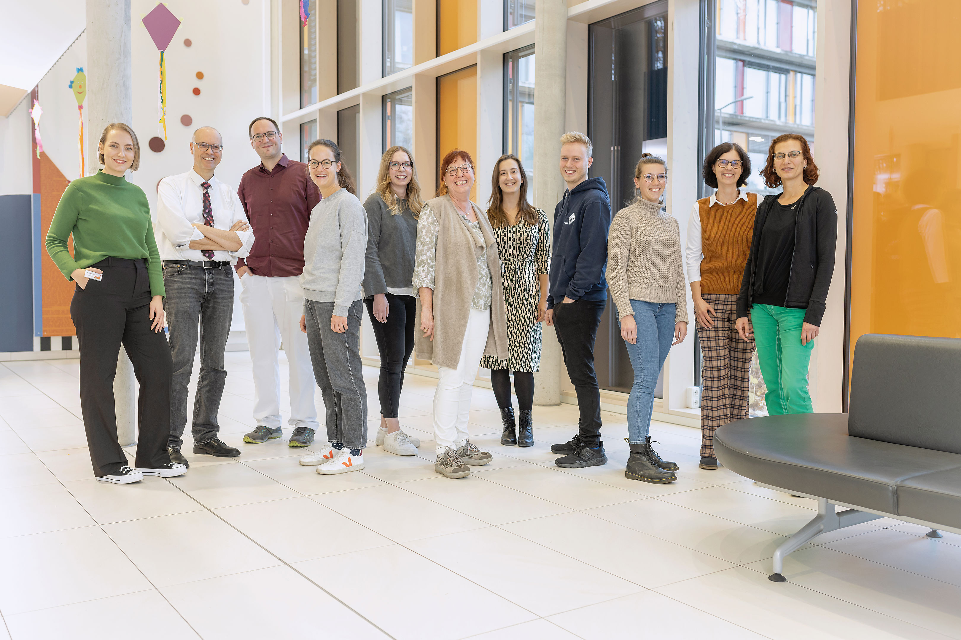 Eine bunt gemischte Gruppe von 12 lächelnden Menschen steht in einem modernen Innenraum mit großen Fenstern und farbenfrohem Dekor.