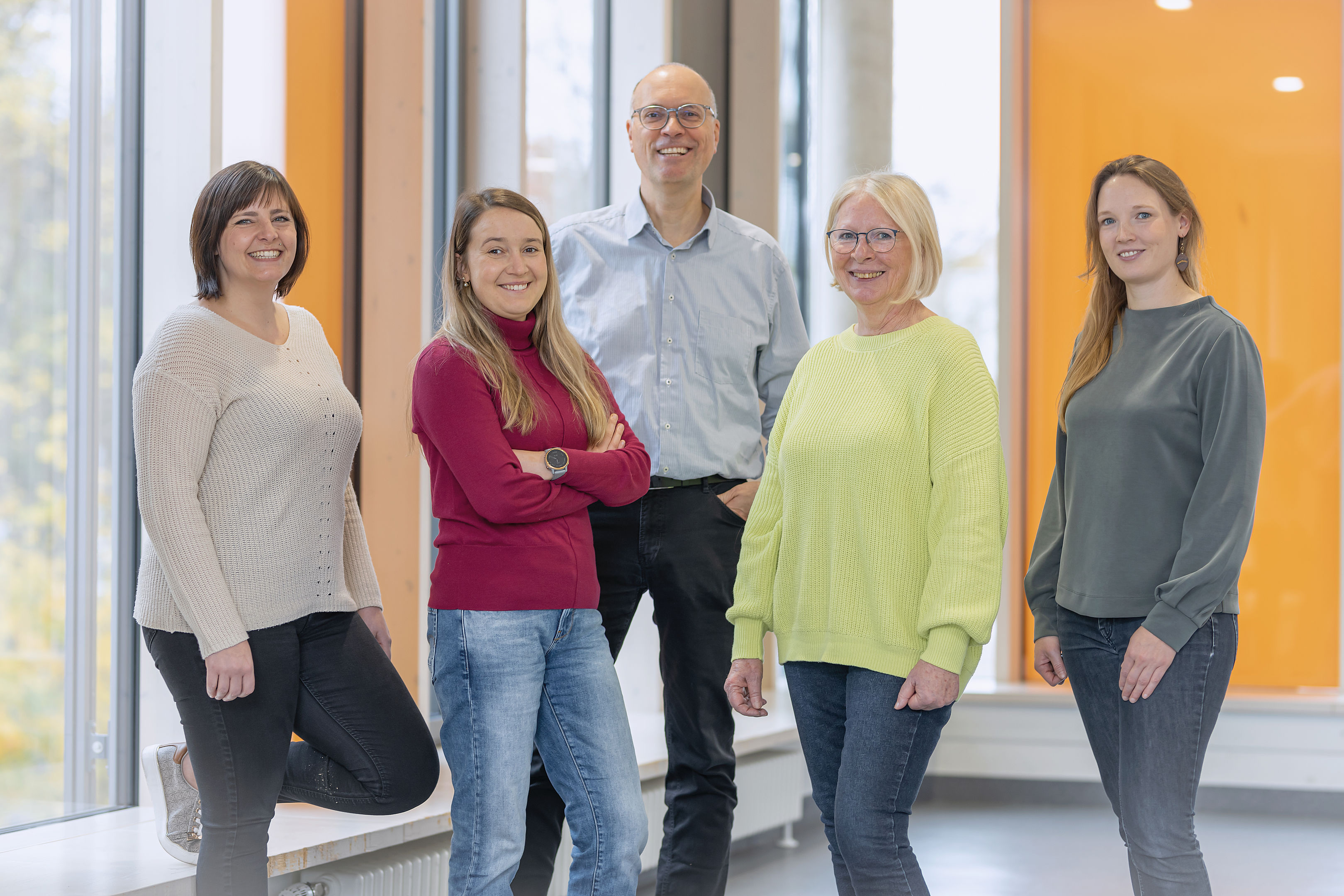 Fünf Menschen stehen in einem hell erleuchteten Raum mit großen Fenstern und orangefarbenen Wänden und lächeln in die Kamera.