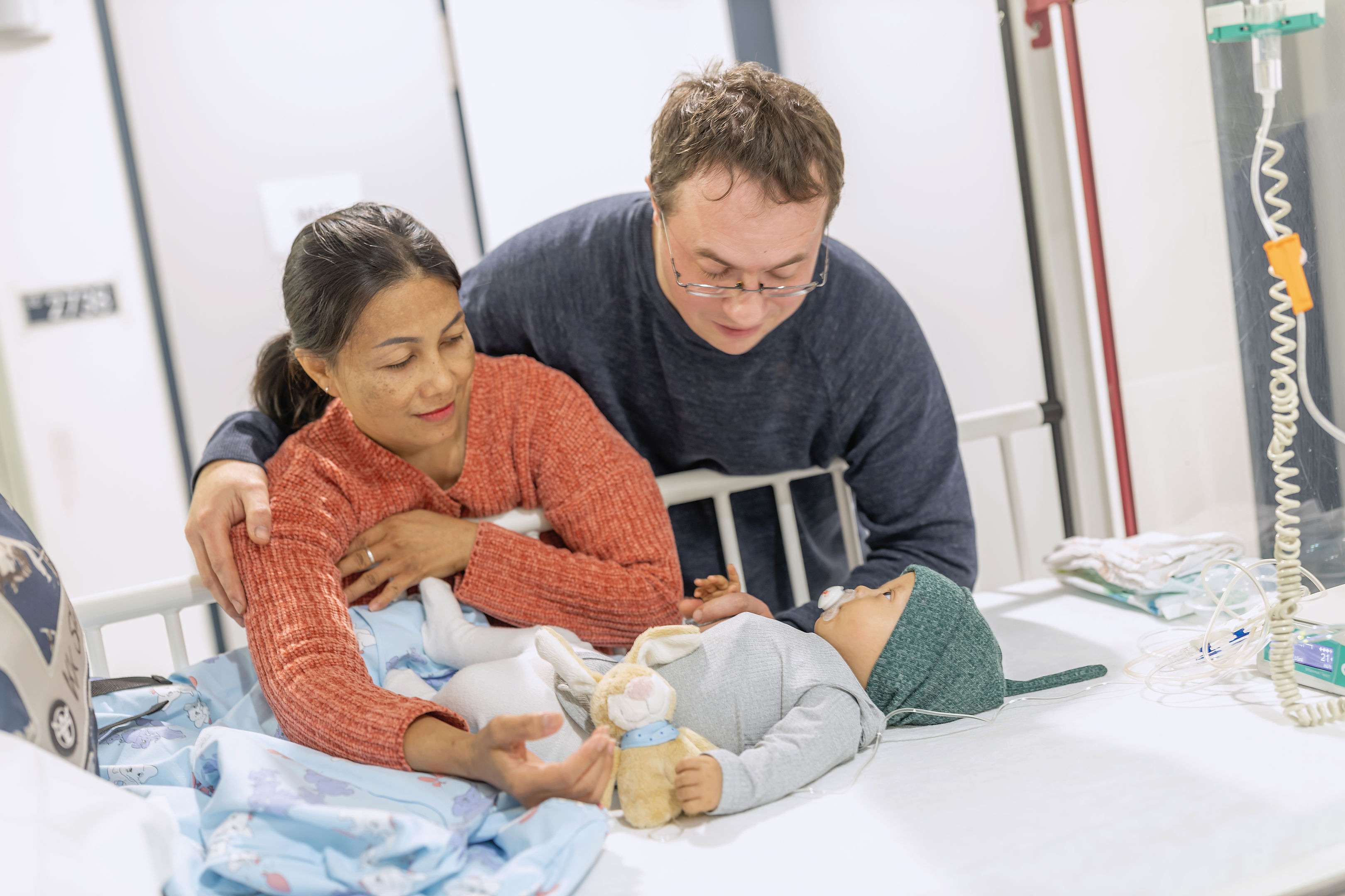 Ein fürsorgliches Paar umarmt ein schlafendes Baby auf einem Krankenhausbett, umgeben von medizinischer Ausrüstung und einem Spielzeug.