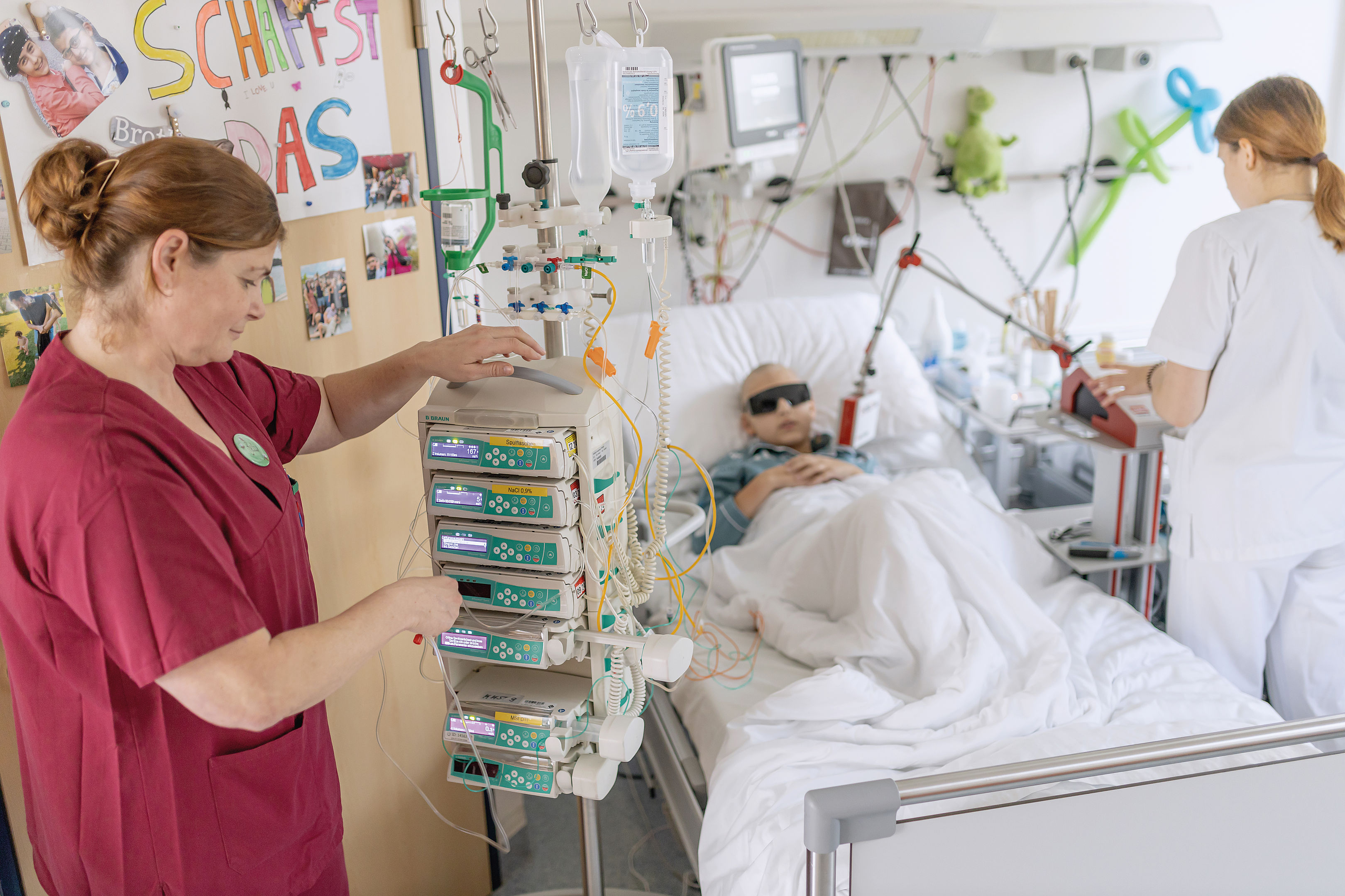 Zwei Krankenschwestern kümmern sich um einen Patienten in einem Krankenzimmer mit medizinischen Geräten und Familienfotos an der Wand.