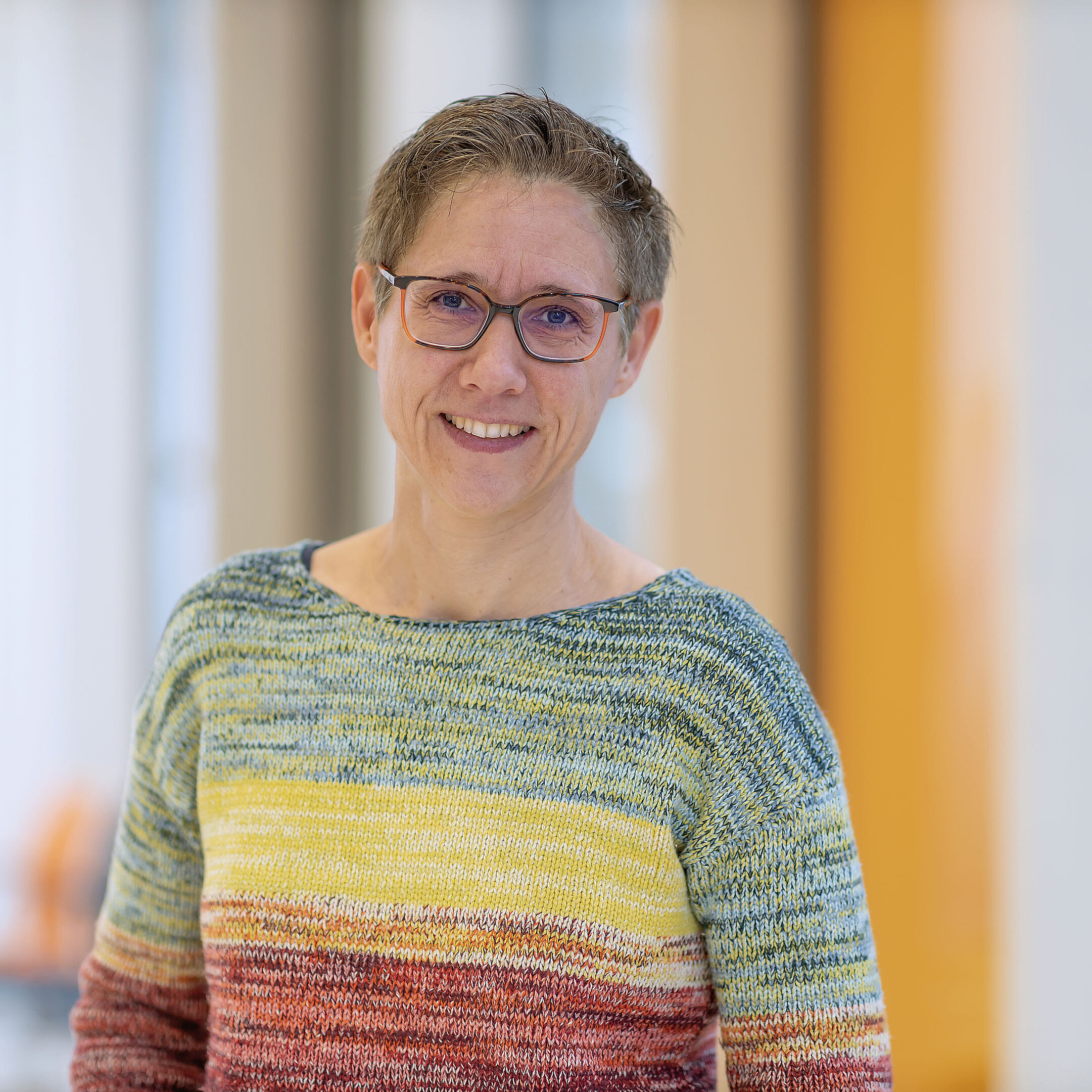 Michaela Kuhnen_Frau mit Brille und buntem, gestreiftem Pullover, lächelt im Innenbereich vor unscharfem Hintergrund.
