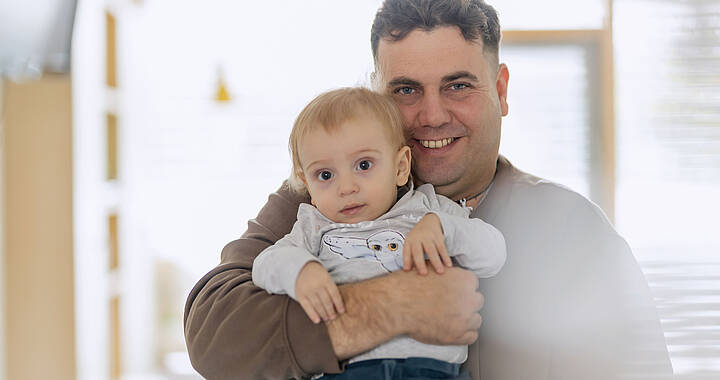 Ein Mann lächelt, während er im Haus ein kleines Kind hält. Beide blicken in die Kamera.