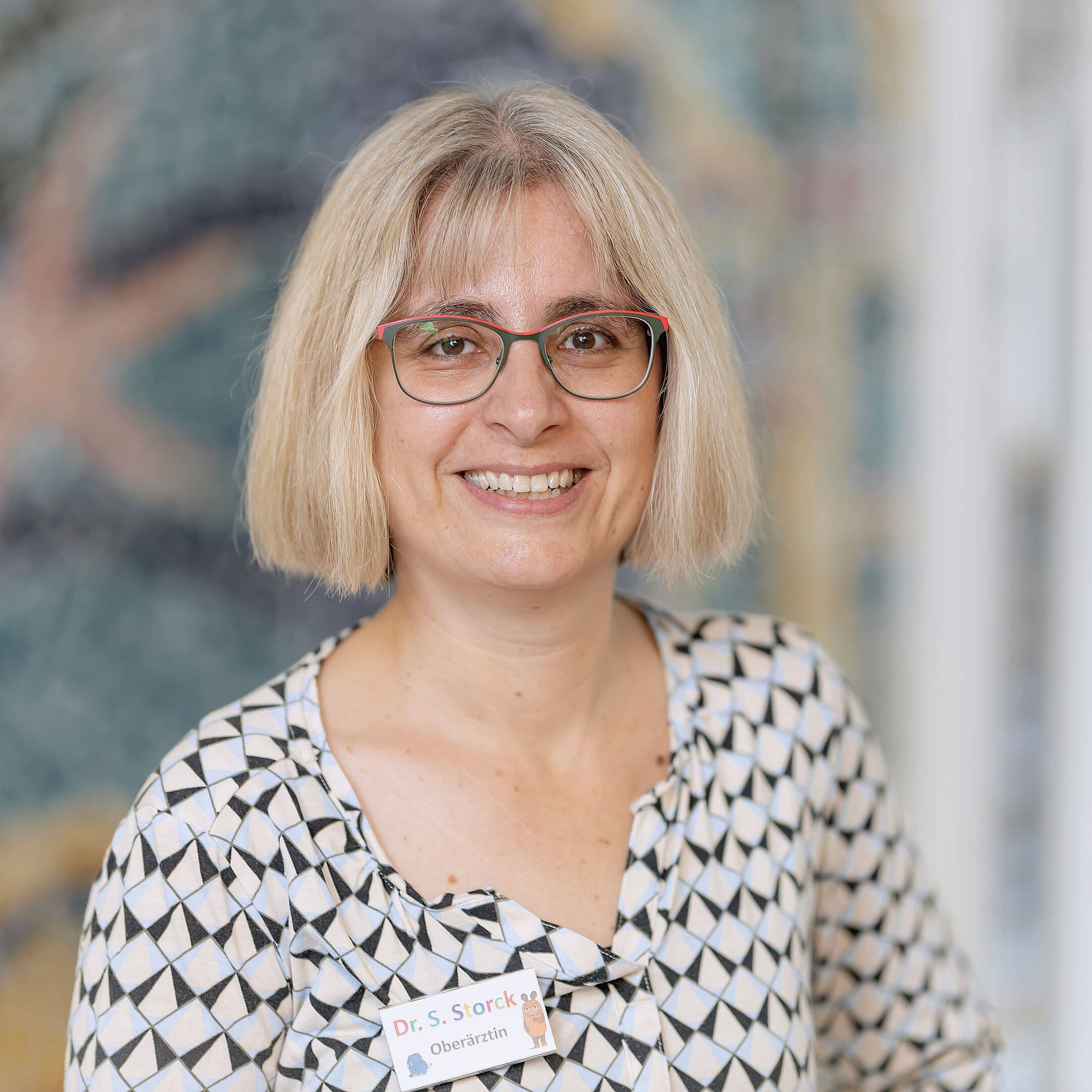 Eine lächelnde Frau mit kurzen blonden Haaren und Brille sowie einer gemusterten Bluse steht im Haus und hält einen Ausweis in der Hand.