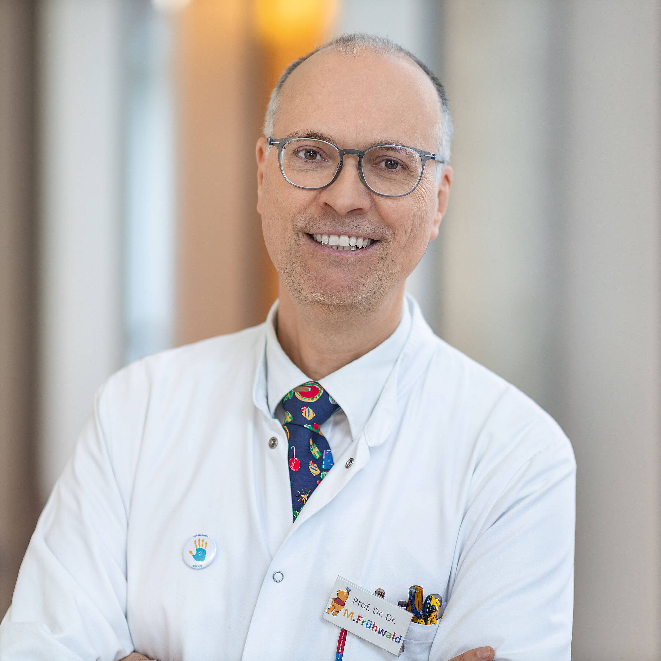 Ein lächelnder Arzt im weißen Kittel steht mit verschränkten Armen in einem gut beleuchteten Raum.