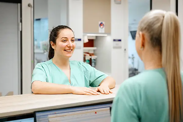 Eine Pflegekraft in grüner Arbeitskleidung unterhält sich an einem Empfangstresen mit einer Kollegin.