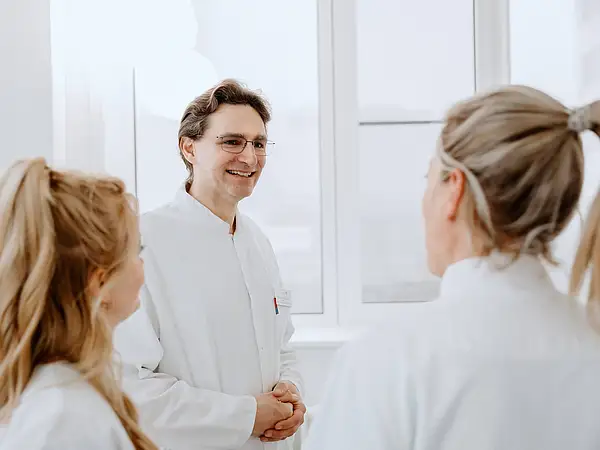 Ein Arzt steht im Gespräch mit zwei Pflegekräften, die ihm aufmerksam zuhören. Alle tragen weiße Arbeitskleidung und befinden sich in einem hellen, modernen Raum.