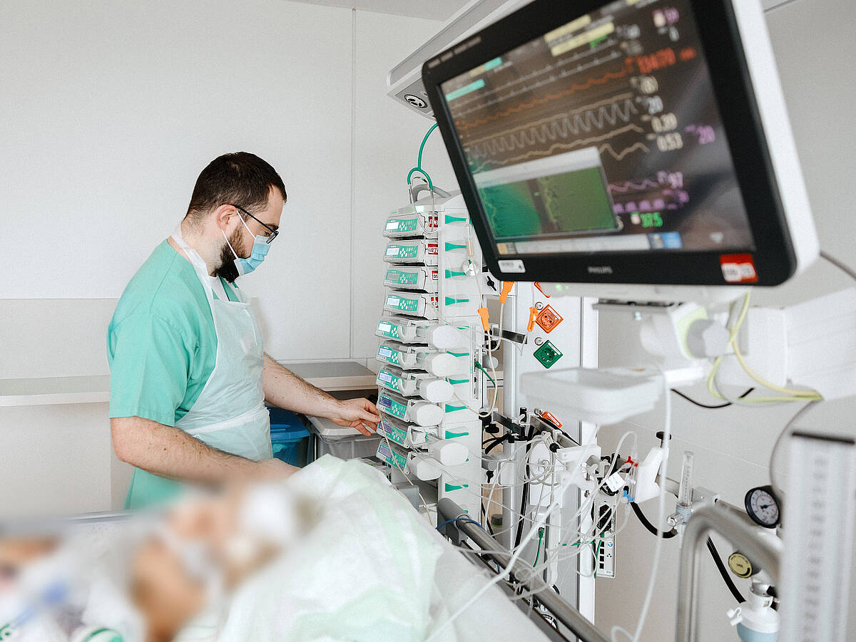 Ein Pflegekraft in grüner Arbeitskleidung stellt ein medizinisches Gerät in einem Krankenhauszimmer ein. Ein Patient ist im Hintergrund zu sehen, während die Pflegekraft den Monitor des Geräts bedient.