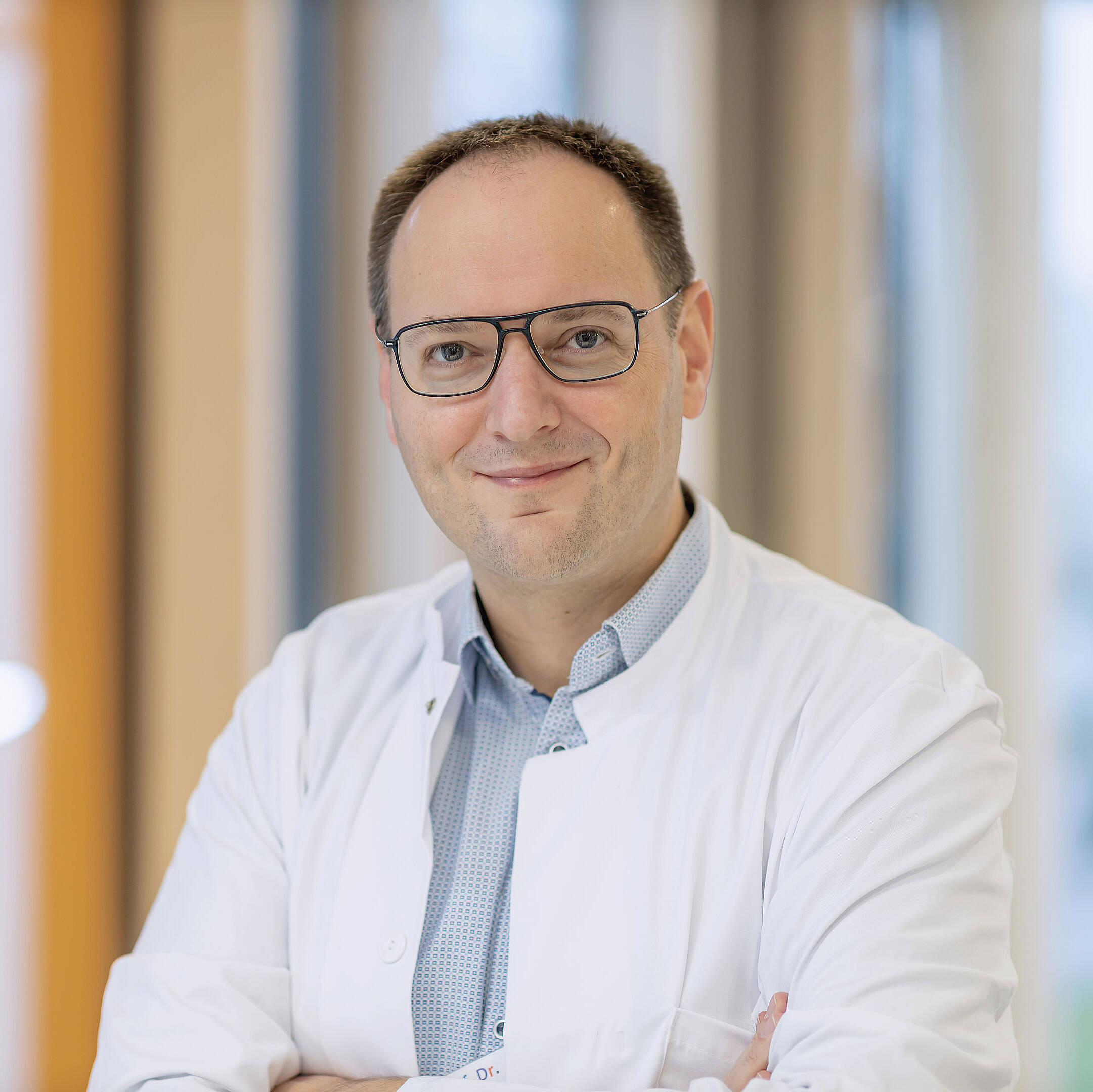 Herr Pascal Johann mit Brille und weißem Kittel, lächelt mit verschränkten Armen in einer hellen, modernen Umgebung.