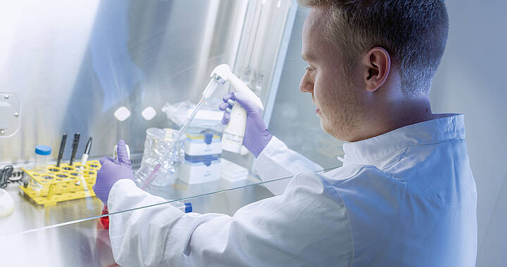 Ein Wissenschaftler im Laborkittel und mit Handschuhen verwendet eine Pipette in einer Laborumgebung, in der Reagenzgläser und Geräte herumstehen.