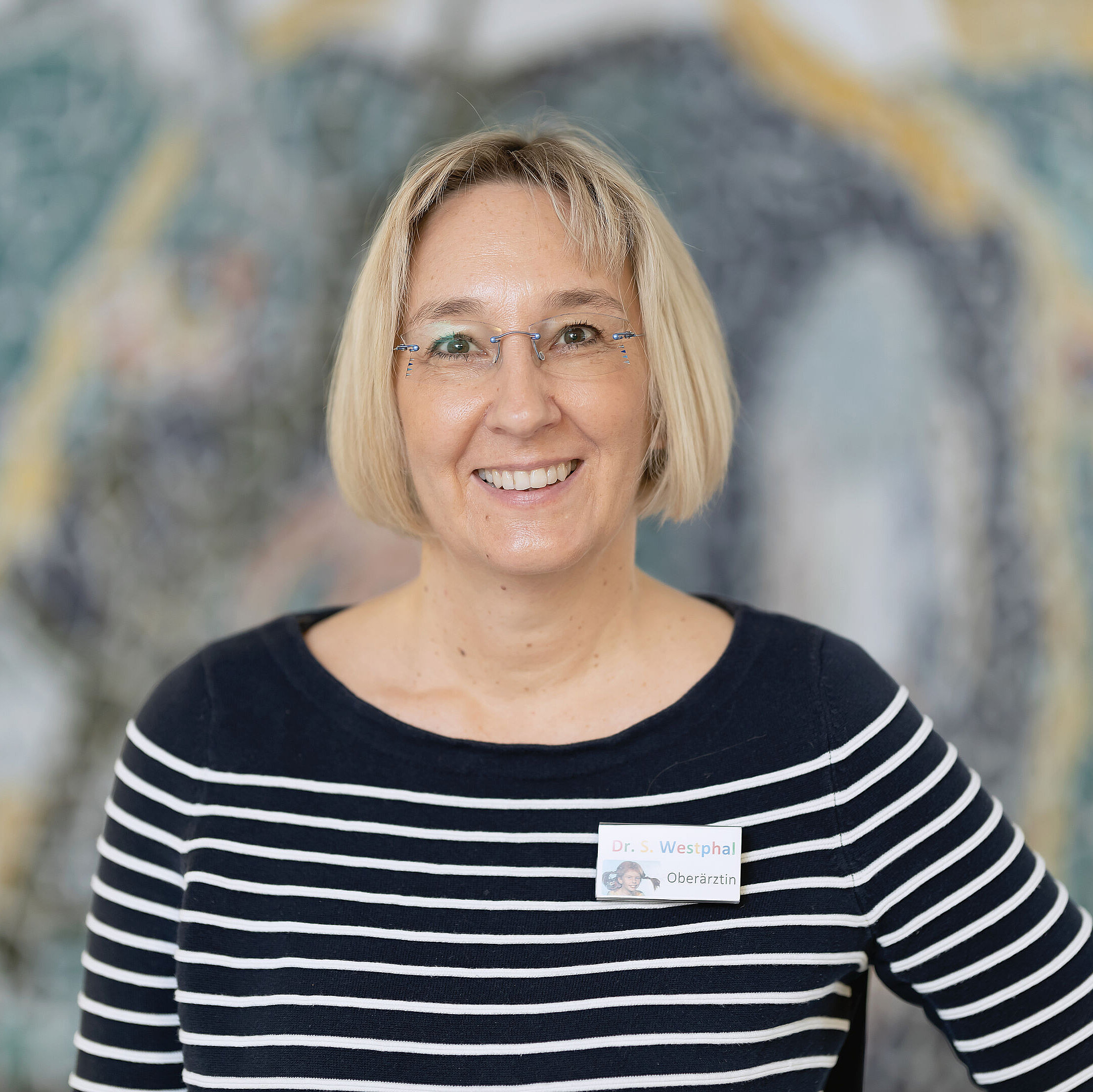 Eine Dr. Silke Westphal mit kurzen blonden Haaren und Brille, einem gestreiften Hemd und einem Namensschild lächelt vor einem verschwommenen Wandgemälde.