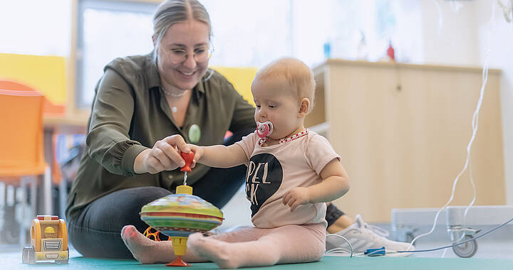 Ein lächelnder Erwachsener und ein Kleinkind spielen in einem hellen Raum mit einem Kreisel auf einer Matte. Das Kleinkind hat einen Schnuller und trägt Rosa.