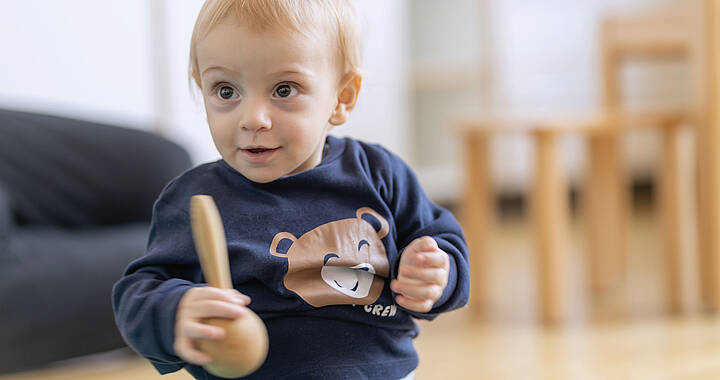 Ein Kleinkind in einem marineblauen Hemd mit Bärenmotiv hält ein Holzspielzeug, während es auf einem Holzboden sitzt. Dahinter sind verschwommene Möbel zu sehen.