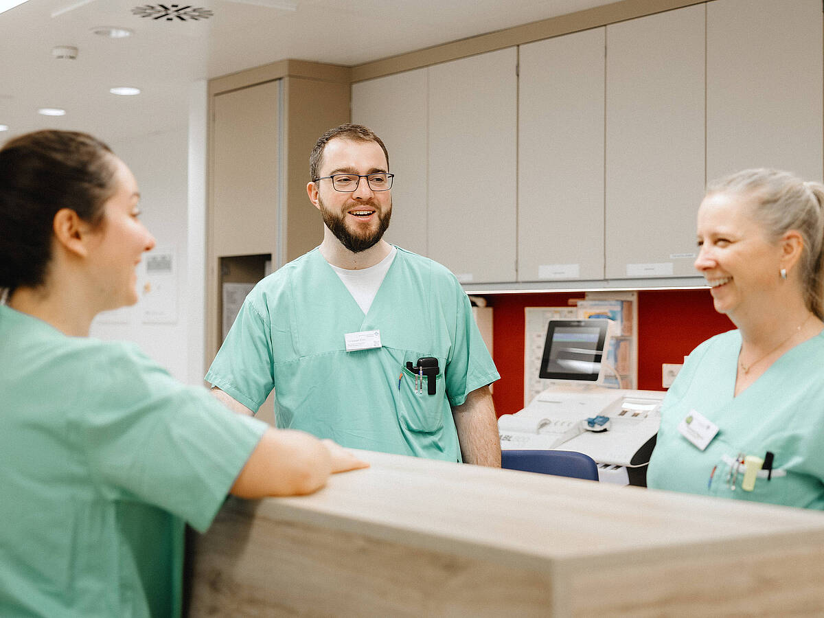 Drei Pflegekräfte in grüner Arbeitskleidung unterhalten sich an einem Empfangstresen in einem Krankenhaus.
