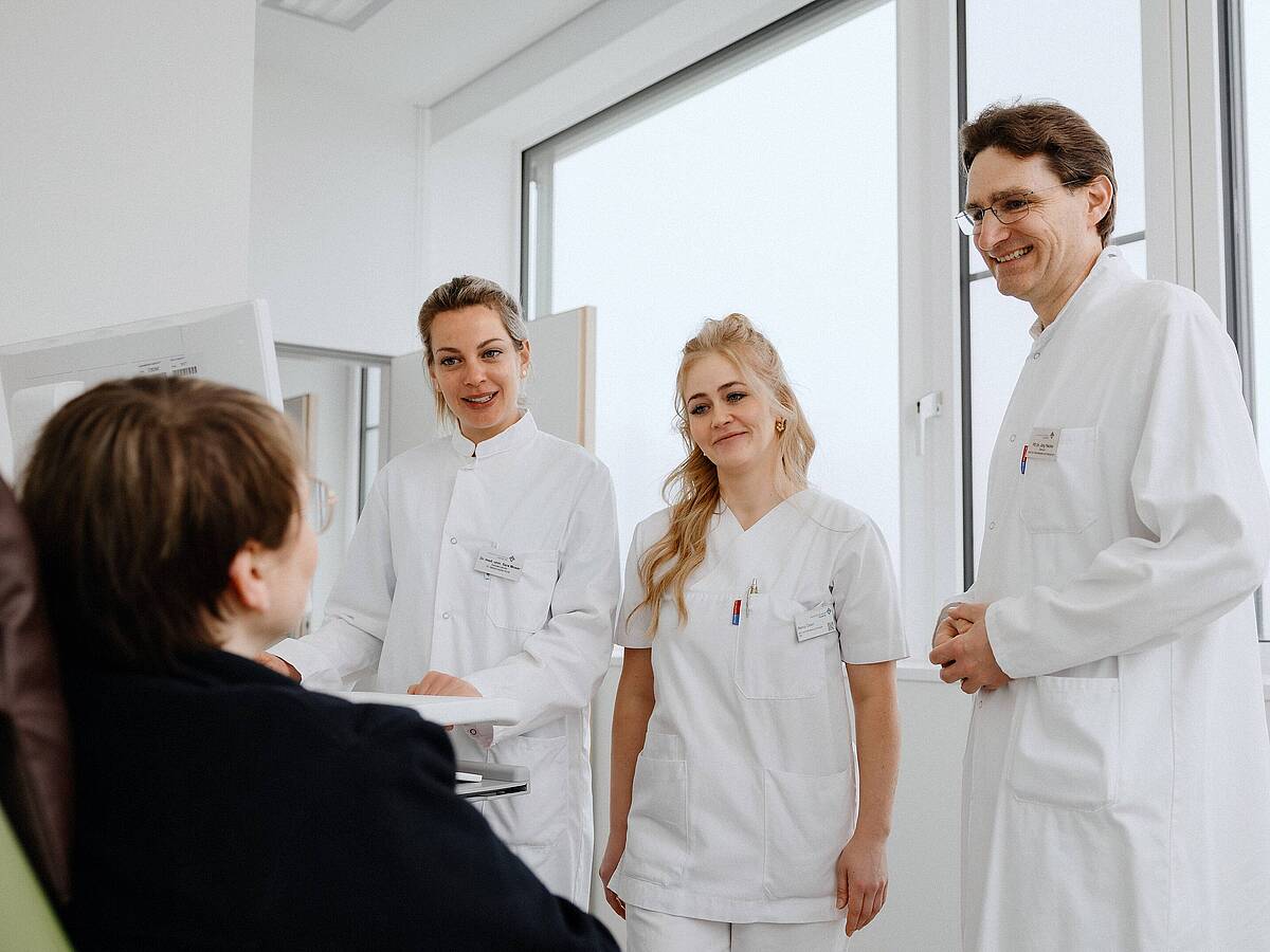 Drei medizinische Fachkräfte in weißen Kitteln unterhalten sich mit einer Patientin in einem Krankenhauszimmer.