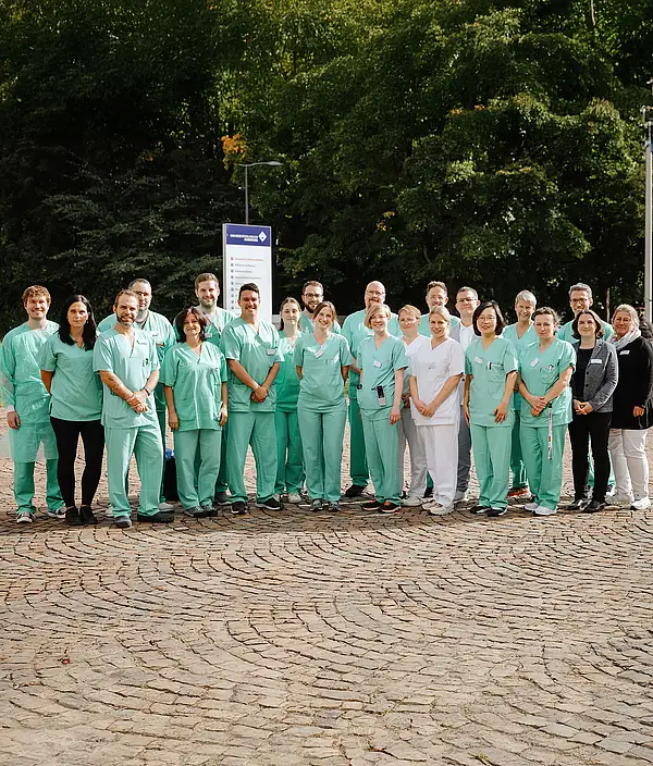 Gruppe von Mitarbeitenden eines Klinikteams in grüner OP-Kleidung steht gemeinsam im Außenbereich vor dem Klinikgelände.