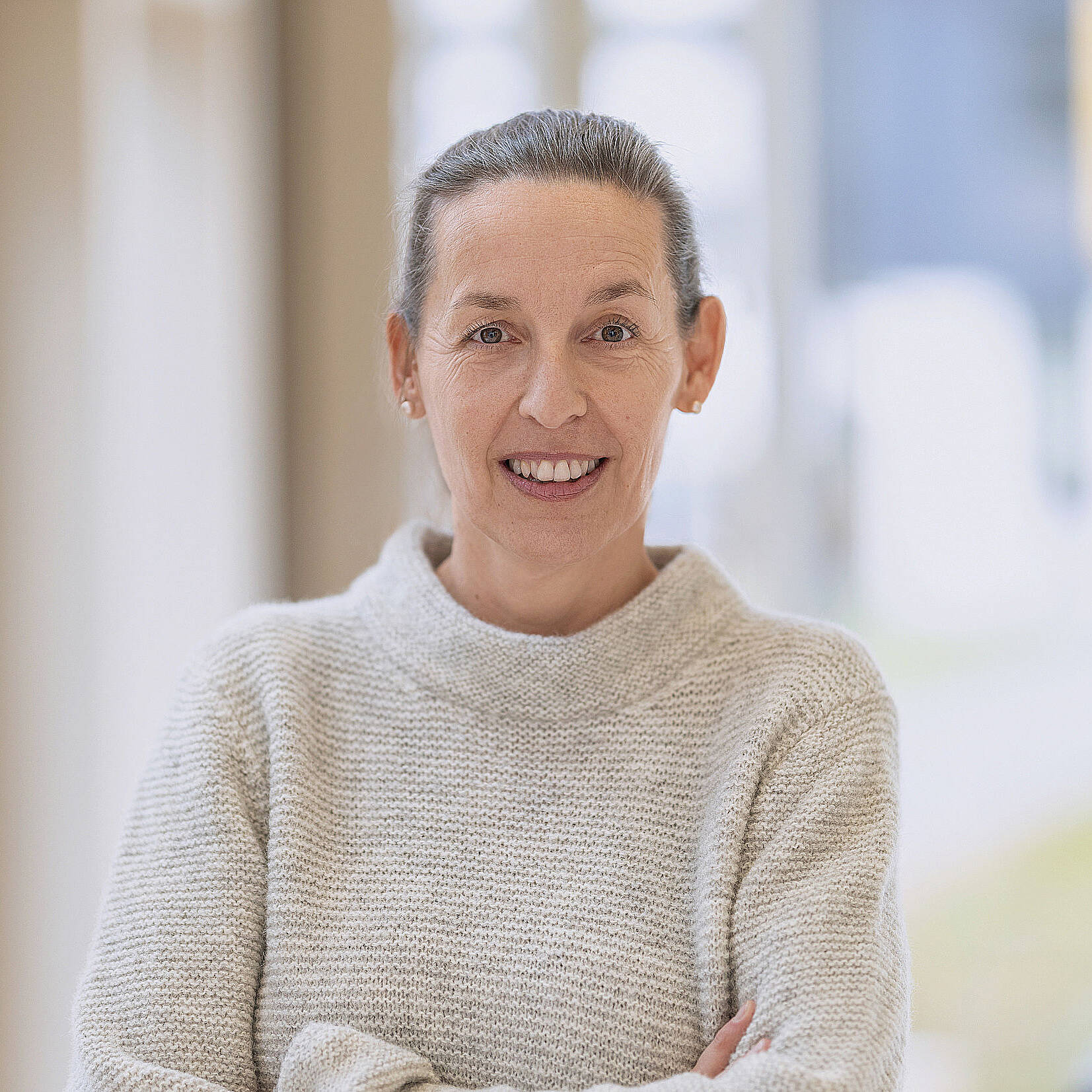 Frau Sonja Ickinger in einem leichten Pullover steht mit verschränkten Armen im Haus und lächelt in die Kamera.