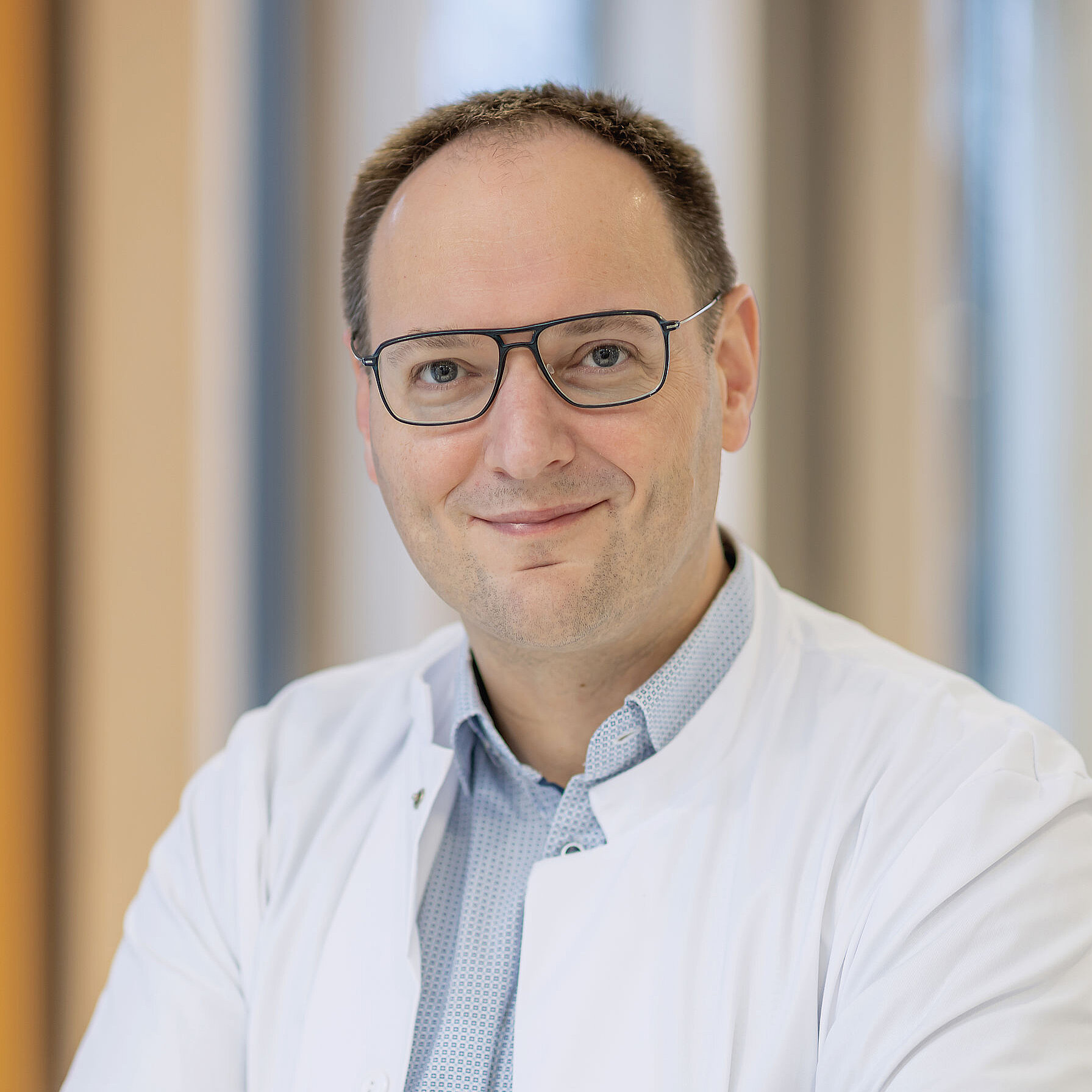 Herr Pascal Johann mit Brille und weißem Kittel, lächelt mit verschränkten Armen in einer hellen, modernen Umgebung.
