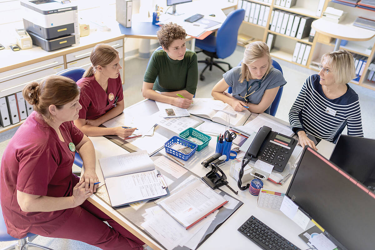 Fünf Personen halten eine Besprechung an einem Bürotisch mit Dokumenten, Computern und Büromaterial ab.