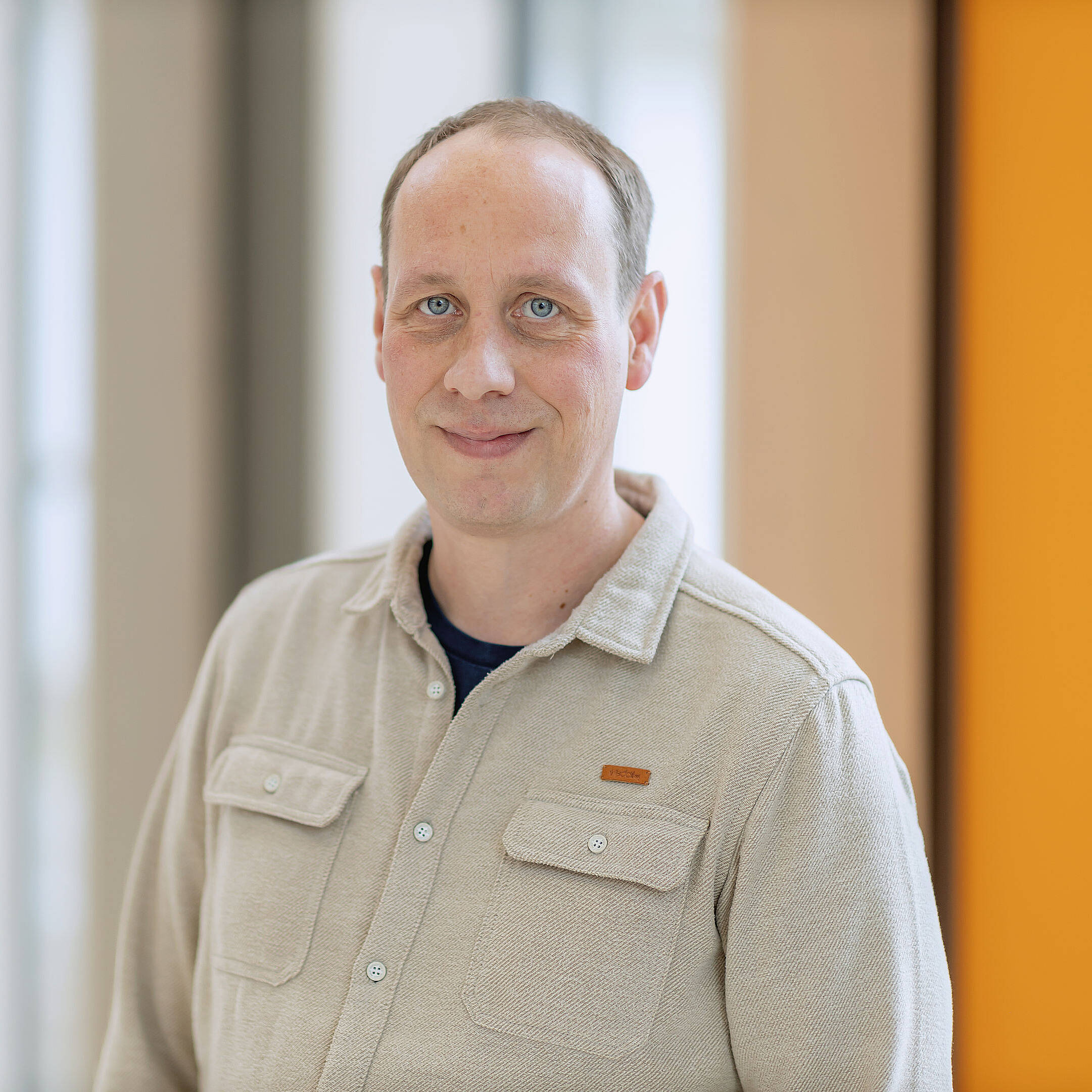 Johannes Holzapfel mit kurzen Haaren in einem beigen Hemd lächelt sanft, während sie im Innenbereich vor einem unscharfen Hintergrund steht.