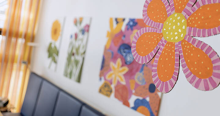 Farbenfrohe Blumenmalereien an einer Wand mit einer Papierblumendekoration im Vordergrund.