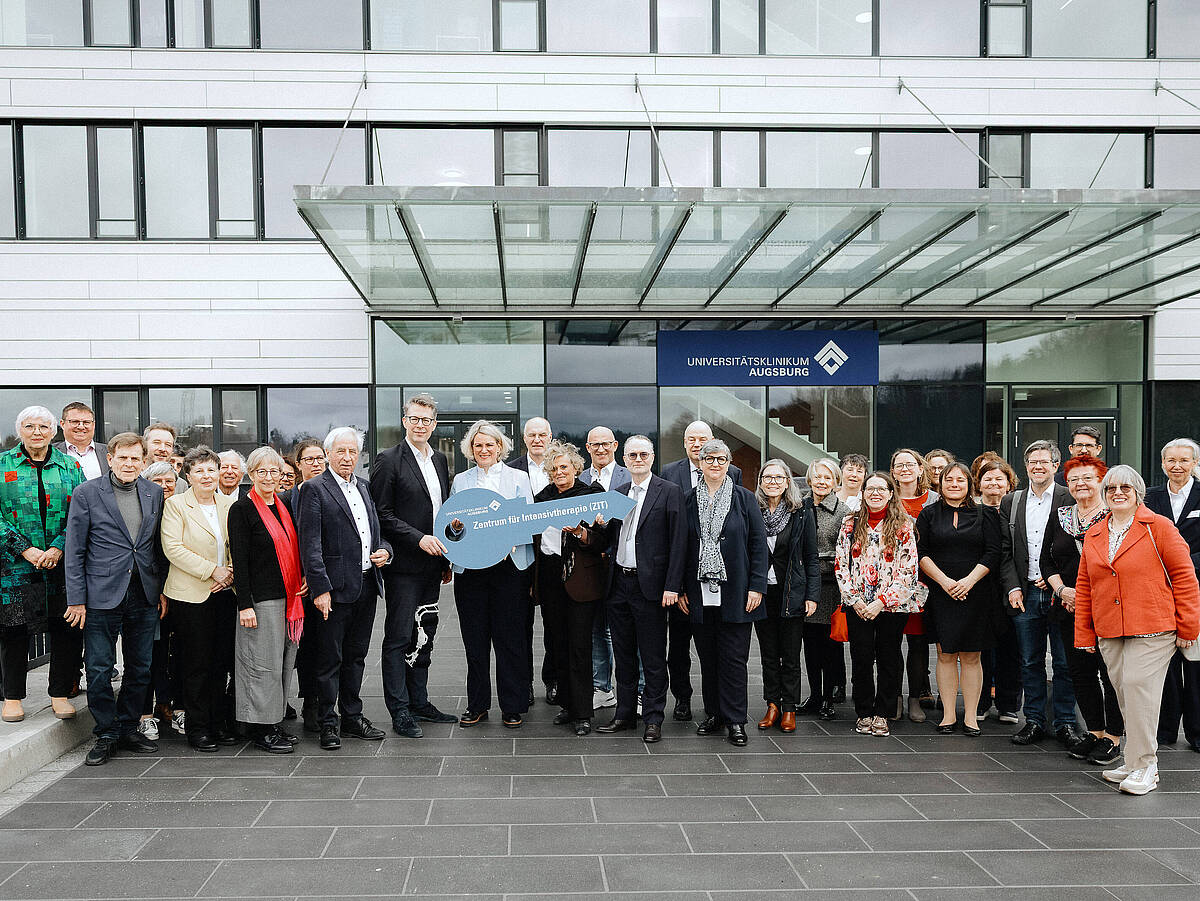 Eine Gruppe von 20 Personen steht vor einem modernen Krankenhausgebäude und posiert für ein Gruppenfoto. Sie tragen unterschiedliche Kleidung, einige in medizinischer Arbeitskleidung und andere in Alltagskleidung.