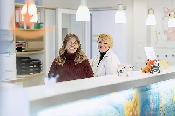 Zwei lächelnde Frauen hinter einem Rezeptionstresen mit lebendigem Dekor und Regalen im Hintergrund.