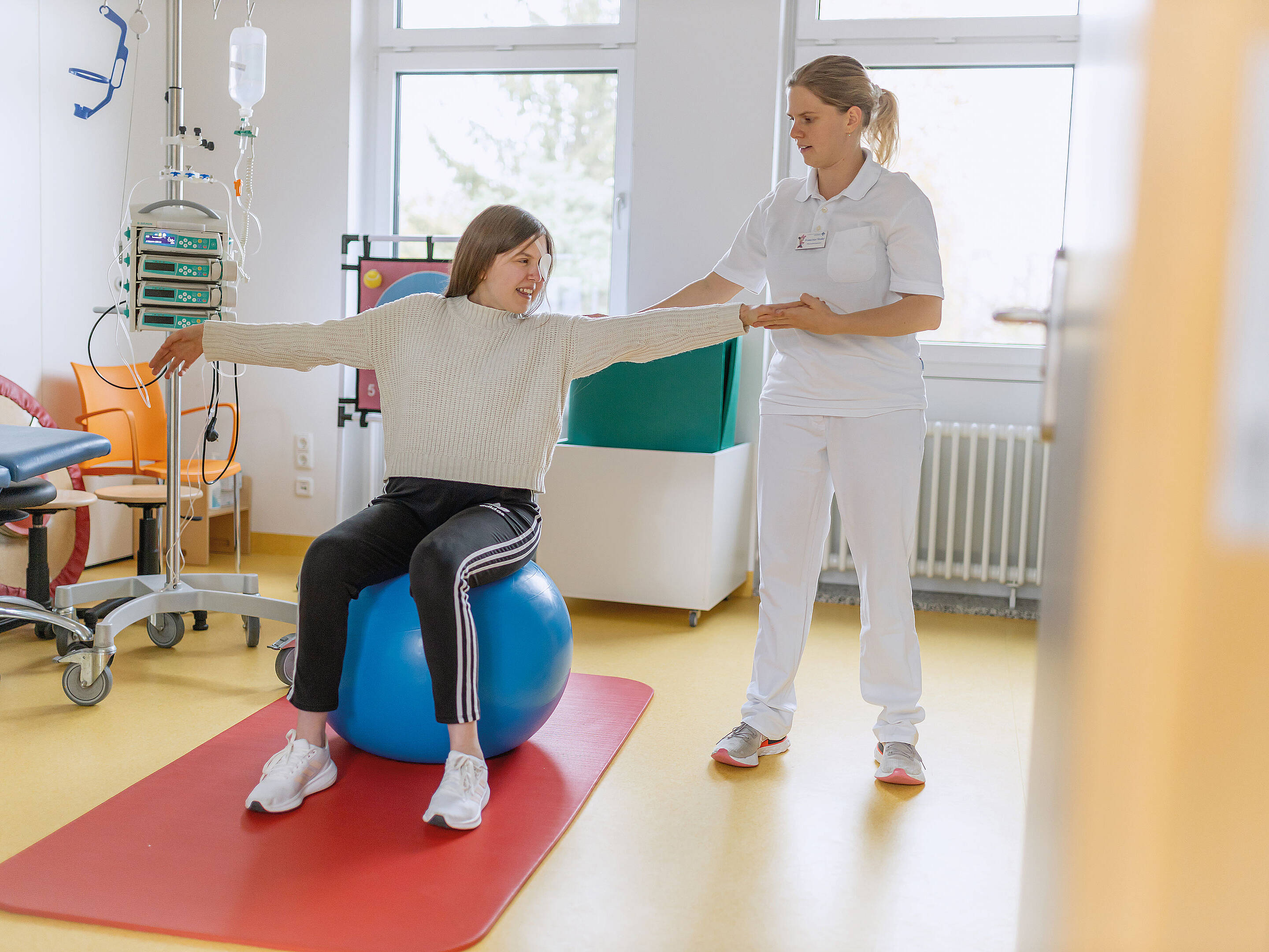 Ein Patient balanciert mit ausgestreckten Armen auf einem Gymnastikball, während ihm in einem hellen Raum eine medizinische Fachkraft Unterstützung bietet.