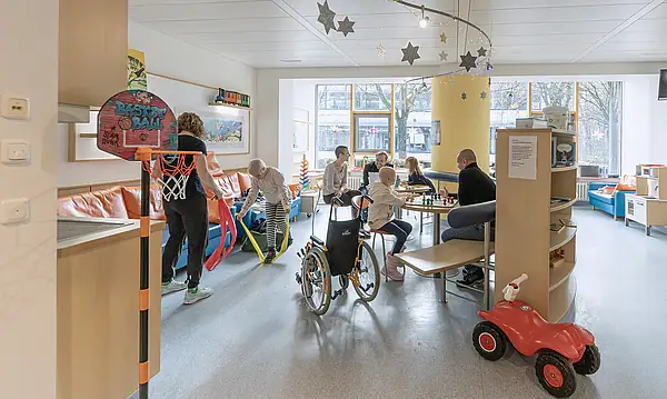 Eine Gruppe von Kindern und Erwachsenen spielt und interagiert in einem hellen Raum mit Spielzeug, einem Basketballkorb und Sternendekorationen.