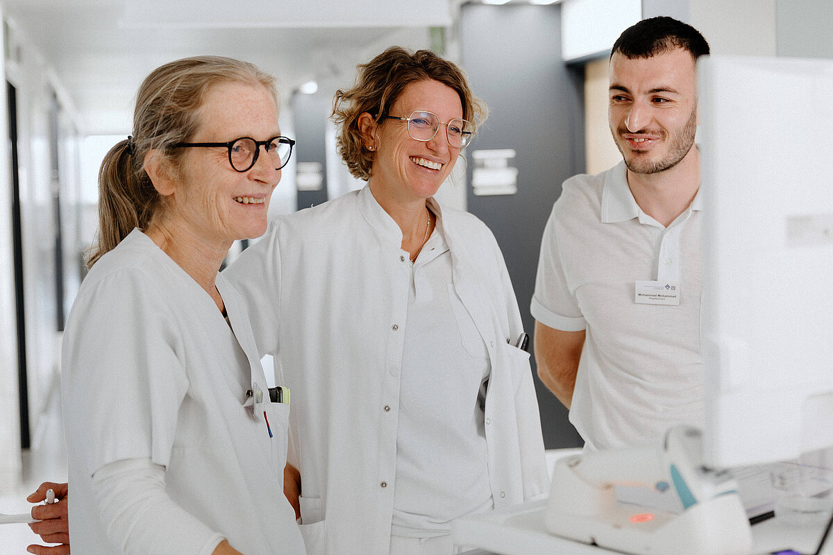 Drei Mitarbeitende im Gesundheitswesen stehen gemeinsam an einem Arbeitsplatz im Krankenhaus und tauschen sich aus.