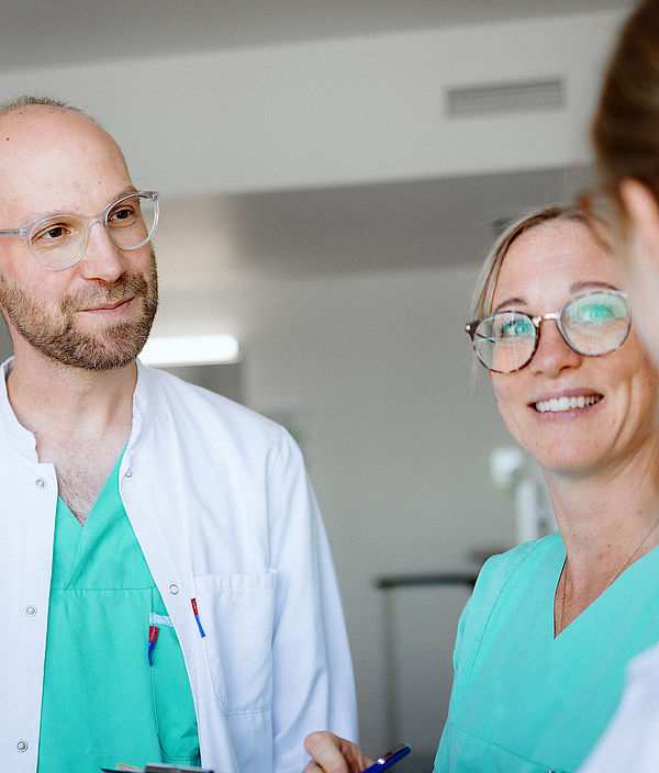 Ein Arzt und eine Pflegekraft im Gespräch, lachen und tauschen sich im Klinikflur aus.