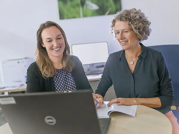 Eine Frau im dunklen Pullover lächelt, während sie sich in einem hellen Raum mit einer anderen sitzenden Frau in einem hellblauen Oberteil unterhält.