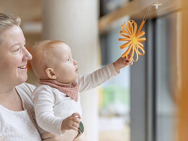 Eine Frau und ein Baby lächeln und greifen nach einer orangefarbenen Papierblume, die an einem Fenster hängt.