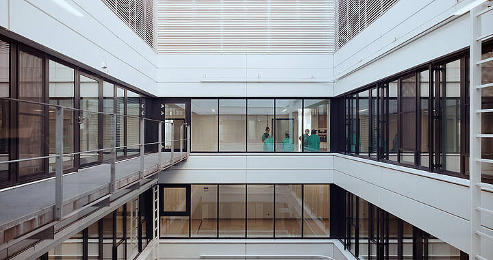 Blick auf das offene Innere eines modernen, mehrstöckigen Gebäudes mit Glaswänden und Metallrahmen. Zwei Personen in grüner Arbeitskleidung sind im Hintergrund zu sehen.