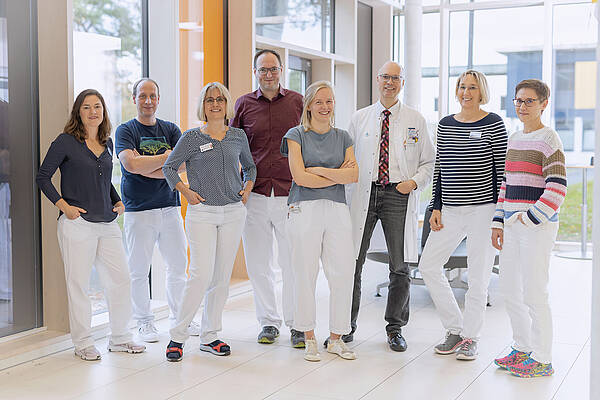 Eine Gruppe von acht Personen, darunter medizinisches Personal, steht lächelnd gemeinsam im Innenbereich an großen Fenstern.