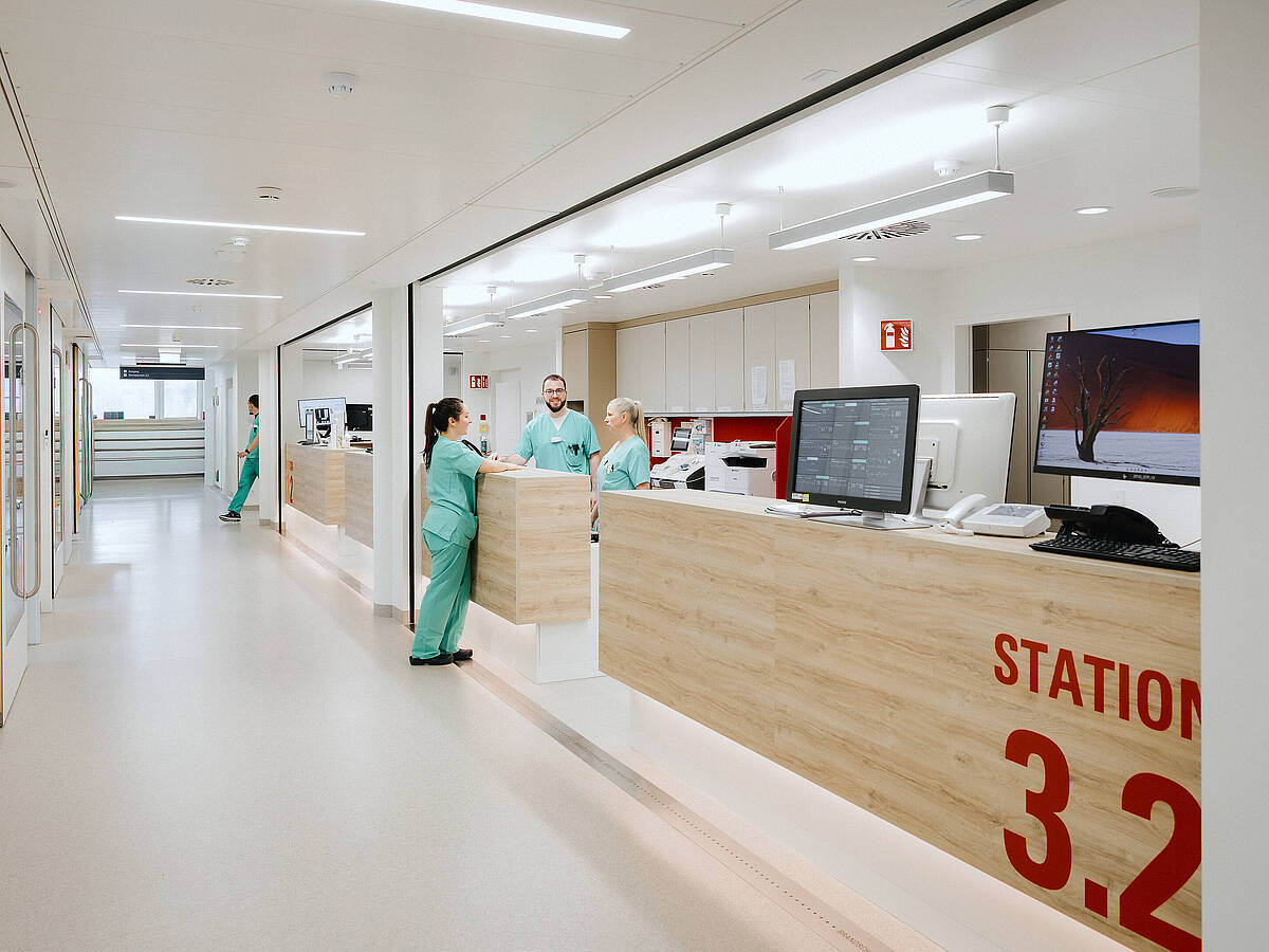 Blick auf den Empfangsbereich einer modernen Krankenhausstation mit einem Empfangstresen. Das Schild „STATION 3.2“ ist sichtbar, und Pflegekräfte arbeiten hinter dem Tresen.