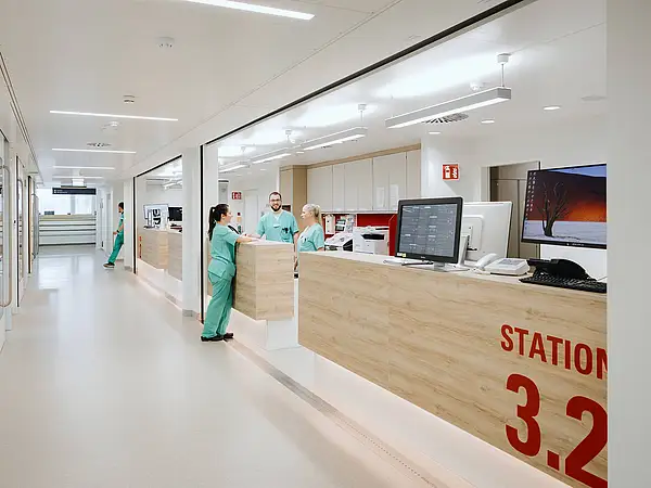 Blick auf den Empfangsbereich einer modernen Krankenhausstation mit einem Empfangstresen. Das Schild „STATION 3.2“ ist sichtbar, und Pflegekräfte arbeiten hinter dem Tresen.