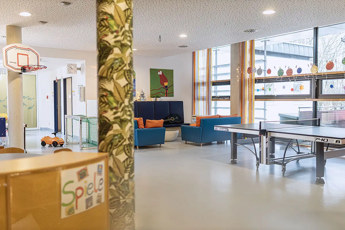 Ein Freizeitraum mit Tischtennisplatte, Basketballkorb, Sofas mit bunten Kissen und großen dekorierten Fenstern.