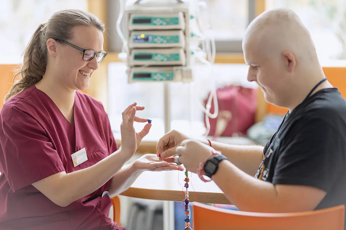 Eine Krankenschwester im kastanienbraunen OP-Kittel hilft einem kahlköpfigen Patienten in einem Krankenhaus, eine Perlenkette anzufertigen.